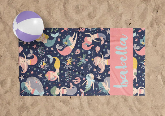 May include: A pink and blue beach towel with a mermaid theme. The towel has a pink stripe on the right side with the name 'Isabella' written in blue. The towel is covered in a pattern of mermaids, stars, and seashells.