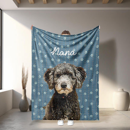 May include: A blue blanket with white paw prints and the name 'Nana' in cursive. The blanket features a large, detailed portrait of a black and gray dog with a red collar. The blanket is held up against a white wall with a window and decorative vases.